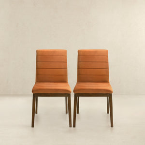The Ines set of 2 modern dining chairs, upholstered in orange with wooden legs, sit side by side against a light beige wall and floor. Their simple design features horizontal seams on the backrest.