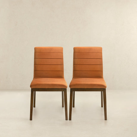 The Ines set of 2 modern dining chairs, upholstered in orange with wooden legs, sit side by side against a light beige wall and floor. Their simple design features horizontal seams on the backrest.