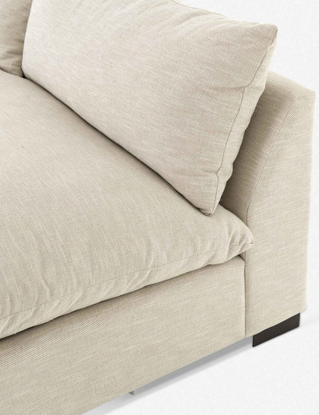Close-up of the Decima Sectional Sofa, featuring beige fabric and a cushioned backrest. The sofa boasts a minimalistic design with straight lines and dark wooden legs, highlighting its cozy and modern aesthetic.