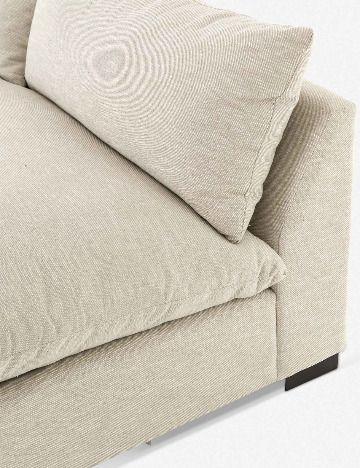 Close-up of the Decima Sectional Sofa, featuring beige fabric and a cushioned backrest. The sofa boasts a minimalistic design with straight lines and dark wooden legs, highlighting its cozy and modern aesthetic.