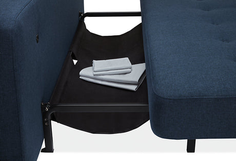 A close-up view of the Deco Convertible Sleeper Sofa, featuring a blue sectional design, reveals a foldable fabric storage compartment between the cushions. Inside the compartment are neatly folded light gray sheets. The sofa is supported by a black metal frame.