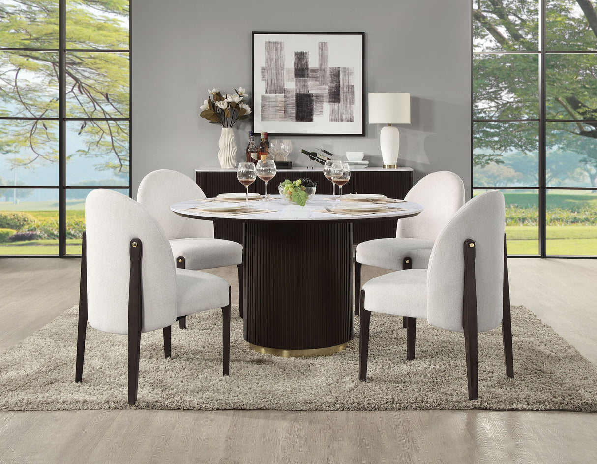 A modern dining room features the Clayten Around Dining Table in marble/espresso, set for four with white upholstered chairs on a textured rug. Large windows showcase a lush view, accompanied by abstract art on minimalist walls and a sideboard adorned with additional decor.