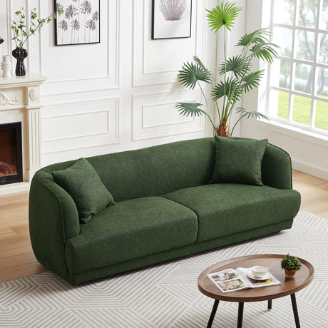 A modern living room showcases the Larisa Mid-Century Modern Linen Sofa in green with two matching cushions. A round wooden coffee table holding books and a plant sits on a light-patterned rug. Decor includes framed wall art, vases on the fireplace mantel, and a potted palm by a large window.