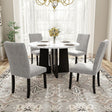 In a bright dining room with large windows and white curtains, a 5 Piece Round Dining Table Set features a white tabletop with a black base. Four gray upholstered chairs surround it, while a vase of yellow flowers is centered on the table, all atop a patterned rug.