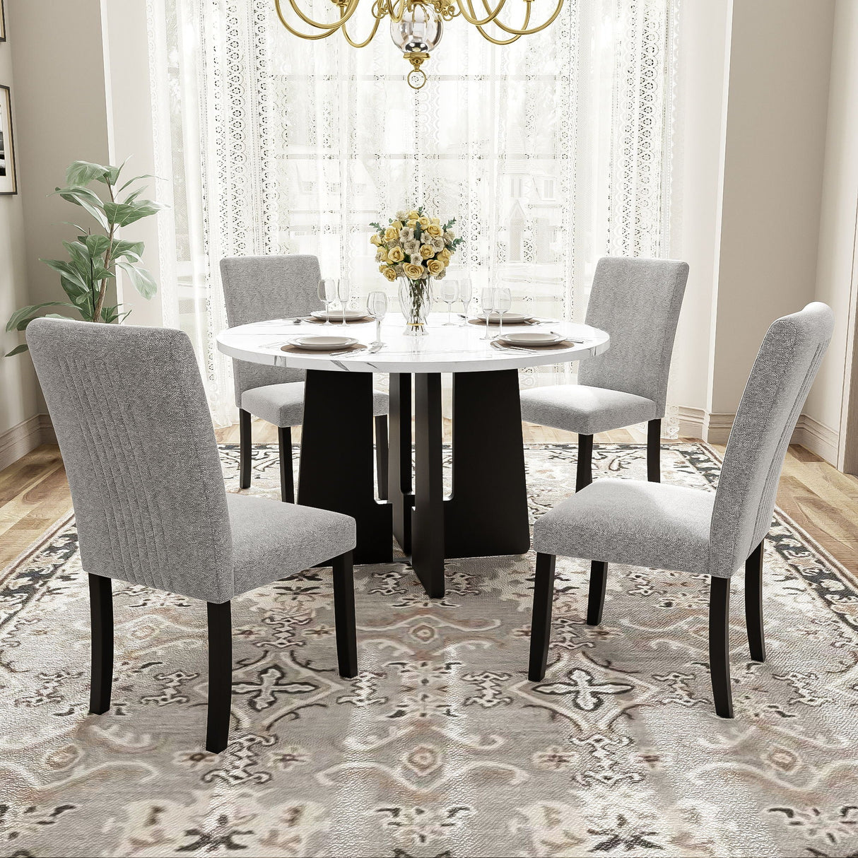 In a bright dining room with large windows and white curtains, a 5 Piece Round Dining Table Set features a white tabletop with a black base. Four gray upholstered chairs surround it, while a vase of yellow flowers is centered on the table, all atop a patterned rug.