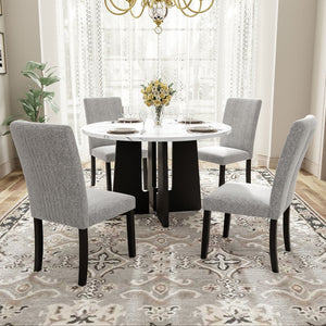 In a bright dining room with large windows and white curtains, a 5 Piece Round Dining Table Set features a white tabletop with a black base. Four gray upholstered chairs surround it, while a vase of yellow flowers is centered on the table, all atop a patterned rug.
