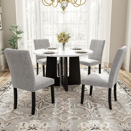 In a bright dining room with large windows and white curtains, a 5 Piece Round Dining Table Set features a white tabletop with a black base. Four gray upholstered chairs surround it, while a vase of yellow flowers is centered on the table, all atop a patterned rug.