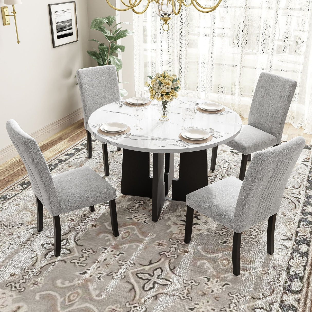 A stylish dining room showcases a 5 Piece Round Dining Table Set with four gray upholstered chairs. The white table is adorned with plates and a vase of yellow flowers, set atop a patterned rug. A luscious plant and lace-curtained window complete the elegant ambiance.