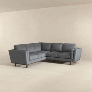 The Erman Pillow Back Corner Sofa in dark gray, featuring an L-shaped design with wooden legs, is set against a plain beige wall. The minimalist room with its light-colored floor highlights the simplicity and elegance of this modern sectional piece.