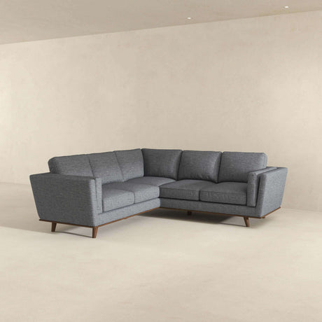 The Erman Pillow Back Corner Sofa in dark gray, featuring an L-shaped design with wooden legs, is set against a plain beige wall. The minimalist room with its light-colored floor highlights the simplicity and elegance of this modern sectional piece.