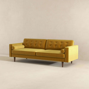 The Casey Mid Century Modern Sofa is a mustard yellow, featuring button-tufted back cushions, cylindrical bolster pillows, and wooden legs. It stands against a neutral-colored wall on a carpeted floor.