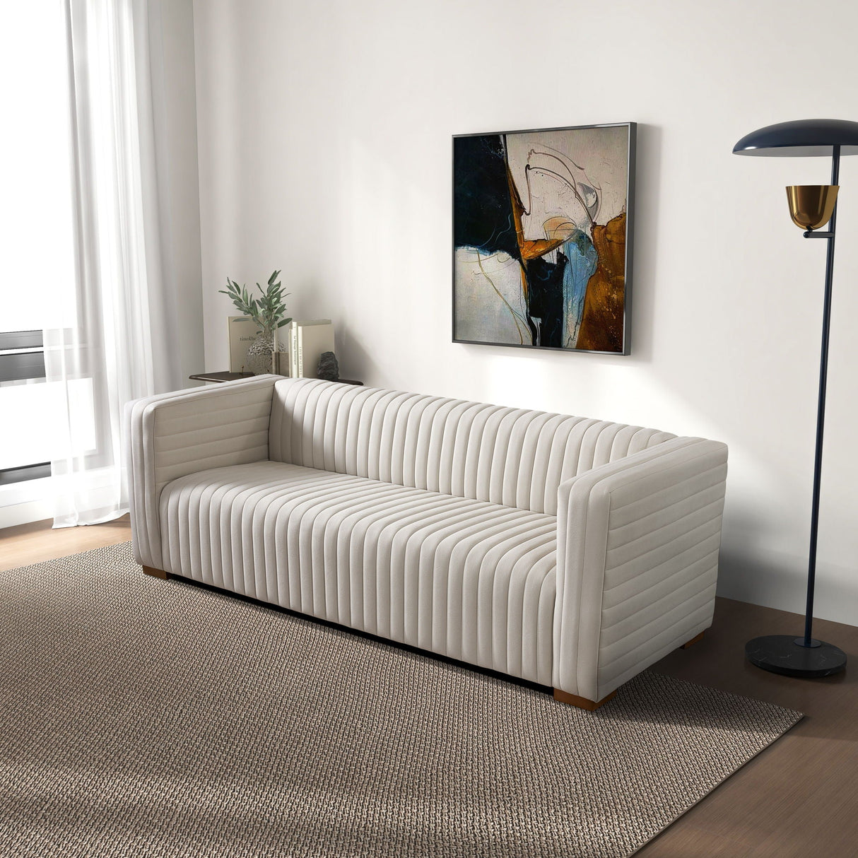A modern living room features the Elara Upholstered Sofa on a textured beige rug. A floor lamp stands beside the sofa. A contemporary abstract painting hangs on the white wall, and a plant sits by the window with sheer curtains.