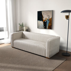 A modern living room features the Elara Upholstered Sofa on a textured beige rug. A floor lamp stands beside the sofa. A contemporary abstract painting hangs on the white wall, and a plant sits by the window with sheer curtains.