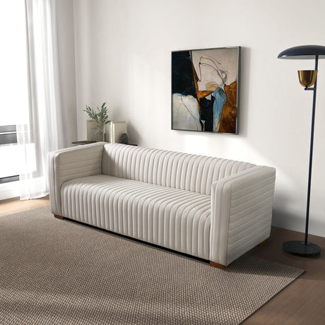 A modern living room features the Elara Upholstered Sofa on a textured beige rug. A floor lamp stands beside the sofa. A contemporary abstract painting hangs on the white wall, and a plant sits by the window with sheer curtains.