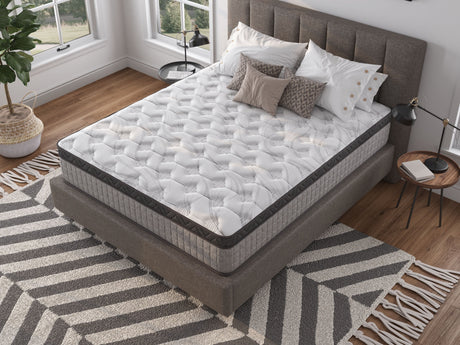 In a modern room, a neatly made bed features the 12" Diamond Innerspring Hybrid and Cooling Gel Memory Foam Mattress. Multiple patterned pillows enhance the look, with a black floor lamp, small round table, large windows, and a zigzag-patterned rug completing the space.