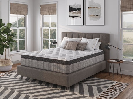 A modern bedroom with a large upholstered bed featuring the 12" Diamond Innerspring Hybrid Cooling Gel Memory Foam Mattress. Accented with decorative pillows, tall windows, abstract wall art, a patterned rug, and a potted plant, it creates a bright, inviting atmosphere.