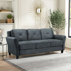 The stylish living room features an iLounge Harvard Microfiber Sofa in dark gray positioned against a white wall. Above the sofa hangs a sunburst mirror, while a potted plant sits on the right side. Built-in shelves with décor adorn the left, and a light-colored rug covers the wooden floor.