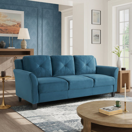 A cozy living room features an iLounge Harvard Microfiber Sofa in teal against a textured blue wall. A lamp and decorative items are arranged on a wooden console, while a round wooden coffee table displays candles and books. Large windows provide ample natural light that fills the space.