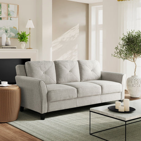 A modern living room with an iLounge Harvard Microfiber Sofa in light gray, a wicker side table, and a glass coffee table adorned with candles. The space is complemented by a potted plant, a table lamp, framed wall art, and large windows allowing ample natural light.