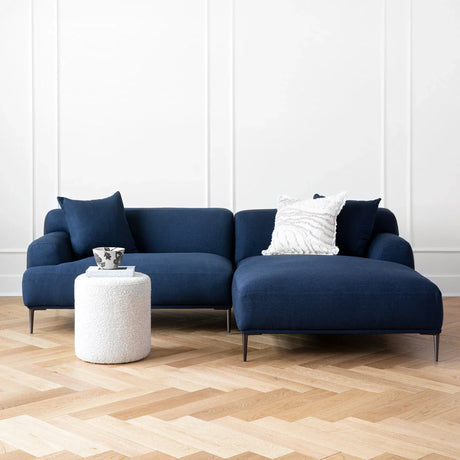 The living room is styled with the Abisko 94.5 Right Sectional in Aurora Blue, which includes two cushions. In front of the sectional, there's a small white textured cylindrical side table featuring a decorative bowl. The space is completed with hardwood flooring and plain white walls.