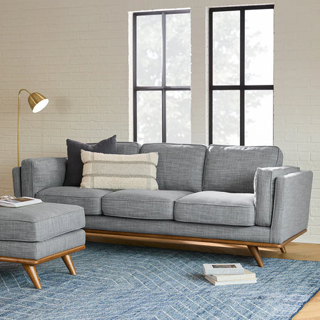 A modern living room featuring the Timber 90" sofa in gray fabric accompanied by a matching ottoman, all set elegantly on a blue patterned rug. The sofa is adorned with two cushions: one in solid gray and the other with stripes. A gold floor lamp stands beside it, while two tall windows allow natural light to flood the space.