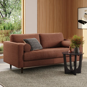 A contemporary living room features the Sven 72 Tufted Loveseat in Ratine Oxide, complemented by two pillows—one rectangular and one cylindrical. A sleek black side table displays a plant and vase. The room is accentuated by wood-paneled walls, a gray textured rug, and a window showcasing leafy green scenery.