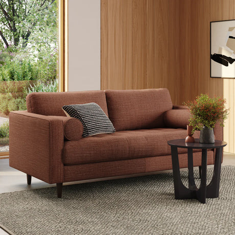 A contemporary living room features the Sven 72 Tufted Loveseat in Ratine Oxide, complemented by two pillows—one rectangular and one cylindrical. A sleek black side table displays a plant and vase. The room is accentuated by wood-paneled walls, a gray textured rug, and a window showcasing leafy green scenery.