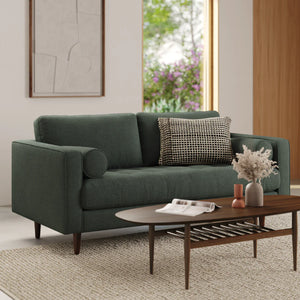 A modern living room featuring the Sven 72 Tufted Loveseat Ratine Oxide, adorned with a dark green cover and paired with a black and white checkered pillow. A wooden coffee table hosts dried flowers and decorative items. The backdrop boasts a large window complemented by abstract wall art.