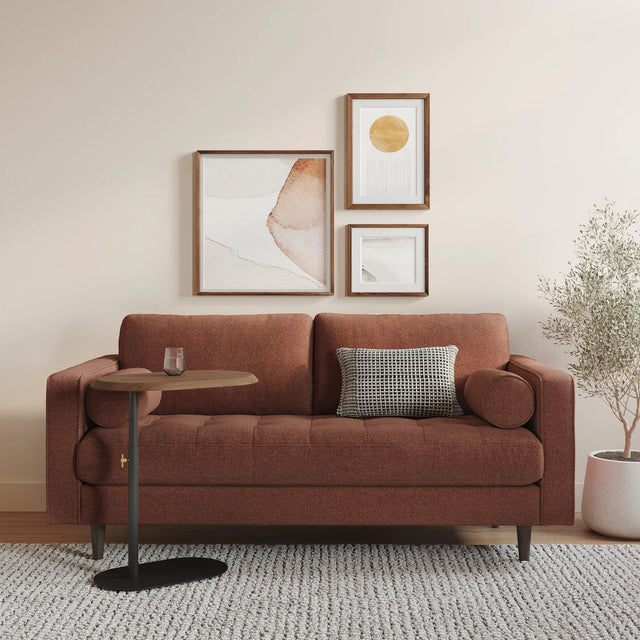 A cozy living room features the Sven 72 Tufted Loveseat in Ratine Oxide, elegantly complemented by two pillows. A small side table holds a glass. Above the loveseat, three abstract framed artworks are displayed on the wall. A potted plant rests on the floor next to a textured rug.