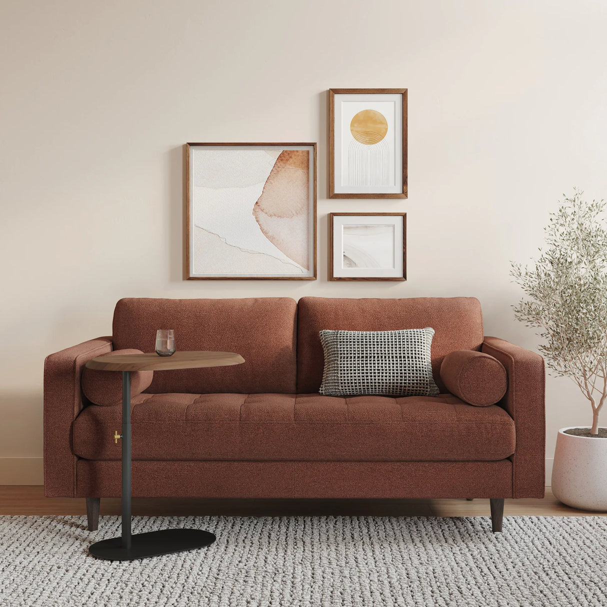 A cozy living room features the Sven 72 Tufted Loveseat in Ratine Oxide, elegantly complemented by two pillows. A small side table holds a glass. Above the loveseat, three abstract framed artworks are displayed on the wall. A potted plant rests on the floor next to a textured rug.