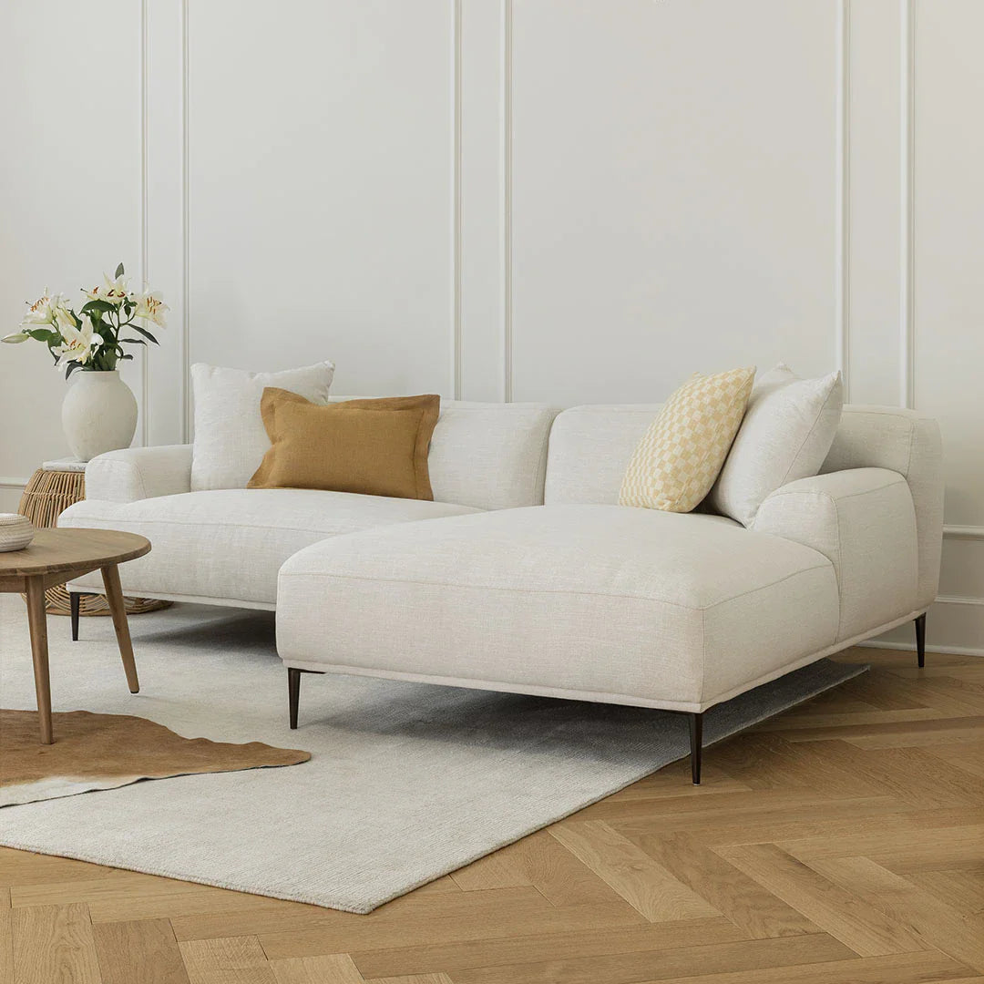 A modern living room is highlighted by the Abisko 94.5 Right Sectional in Aurora Blue, accented with a yellow and white pillow. A vase filled with flowers rests on a small wooden table. The hardwood floor is complemented by a light rug, while the walls are adorned with subtle white panel details.