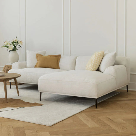 A modern living room is highlighted by the Abisko 94.5 Right Sectional in Aurora Blue, accented with a yellow and white pillow. A vase filled with flowers rests on a small wooden table. The hardwood floor is complemented by a light rug, while the walls are adorned with subtle white panel details.