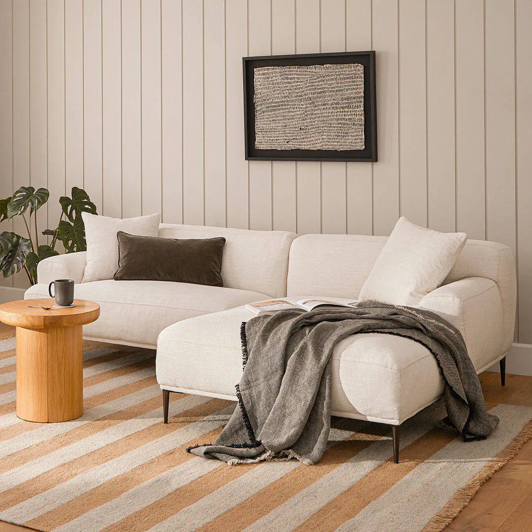 A cozy living room features the Abisko 94.5 Right Sectional in Aurora Blue, topped with a dark throw blanket and pillows. Next to it is a small wooden coffee table with a black mug. A plant rests in the corner, and textured artwork is mounted on the light wood-paneled wall.