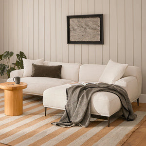 A cozy living room features the Abisko 94.5 Right Sectional in Aurora Blue, topped with a dark throw blanket and pillows. Next to it is a small wooden coffee table with a black mug. A plant rests in the corner, and textured artwork is mounted on the light wood-paneled wall.