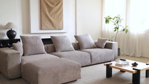A contemporary living room featuring a sizable black 142 in. square arm, 4-piece L-shaped corduroy polyester modern sectional sofa. A wooden coffee table sits in the center, complemented by a black lamp on a side table. A potted plant is placed near the window adorned with sheer curtains, while minimalist artwork graces the wall.