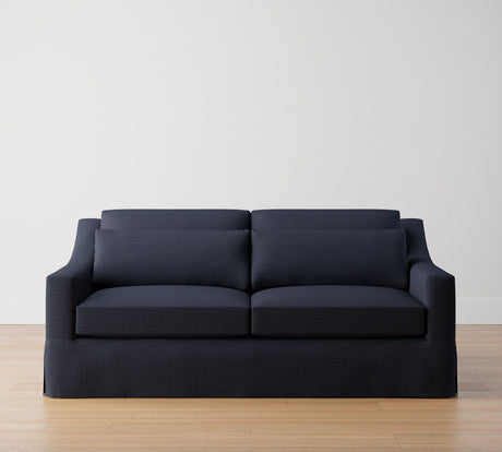 The York Slope Arm Deep Slipcovered Sofa in dark blue, featuring sleek cushions and accommodating two seats, rests on a light wooden floor against a plain white wall. Its straight lines and minimalist design offer a modern and clean aesthetic to the room.