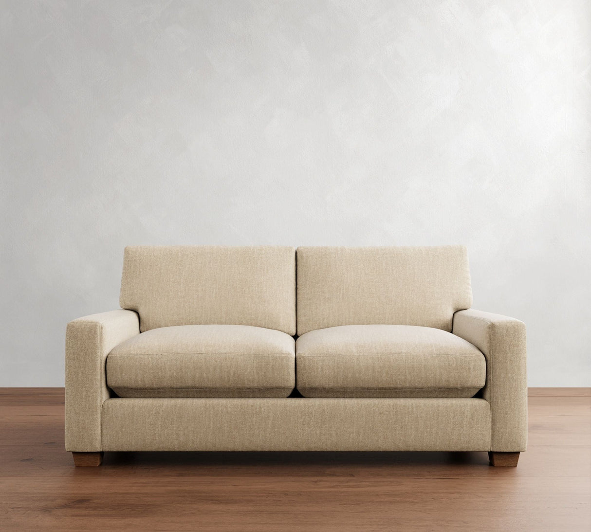 The PB Comfort Modern Square Arm Upholstered Sofa, featuring two seat cushions and two back cushions, is arranged against a light gray textured wall. It sits on a wooden floor and boasts square armrests and wooden legs.