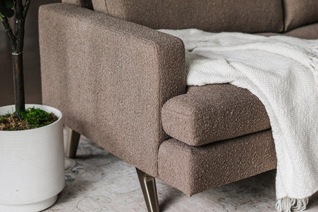 Close-up of the Lido Sofa with a brown textured fabric and sleek metal legs. A soft, white blanket is draped over one arm. A potted plant sits on the floor nearby, adding a touch of greenery to the cozy, modern setting.