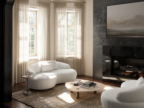 A modern living room features two Rio Sofas in white with a curvy design, paired with a round wooden coffee table and a large painting above a sleek black marble fireplace. Sunlight floods through tall windows draped in sheer curtains, casting light throughout the neutrally-toned space.