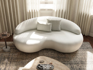A stylish Rio Sofa with a modern, uniquely shaped white design and rounded edges is set against a wall featuring sheer curtains. Three matching cushions are arranged on the sofa. In the foreground, there is a beige rug and a round coffee table adorned with decorative items, all illuminated by natural light filtering in.