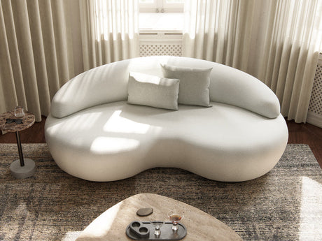 A stylish Rio Sofa with a modern, uniquely shaped white design and rounded edges is set against a wall featuring sheer curtains. Three matching cushions are arranged on the sofa. In the foreground, there is a beige rug and a round coffee table adorned with decorative items, all illuminated by natural light filtering in.