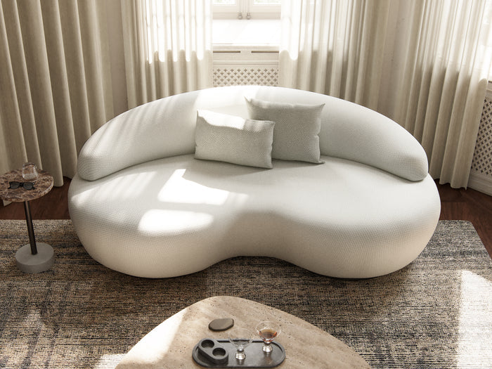 A stylish Rio Sofa with a modern, uniquely shaped white design and rounded edges is set against a wall featuring sheer curtains. Three matching cushions are arranged on the sofa. In the foreground, there is a beige rug and a round coffee table adorned with decorative items, all illuminated by natural light filtering in.