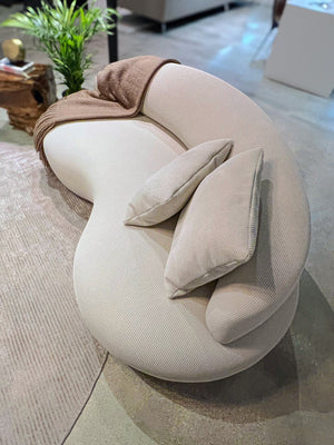 The Rio Sofa, featuring a modern curved design in white, comes with two matching pillows and is elegantly adorned with a brown throw blanket draped over one arm. It's situated on a textured beige rug alongside a small green plant, perfectly complementing the contemporary living room setting.