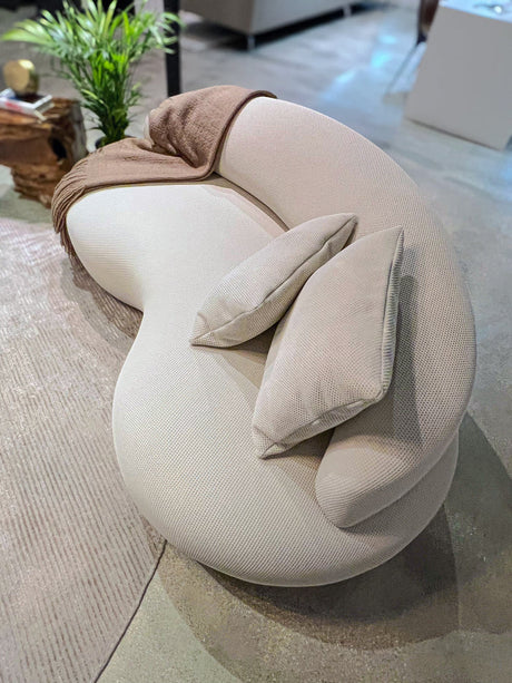 The Rio Sofa, featuring a modern curved design in white, comes with two matching pillows and is elegantly adorned with a brown throw blanket draped over one arm. It's situated on a textured beige rug alongside a small green plant, perfectly complementing the contemporary living room setting.
