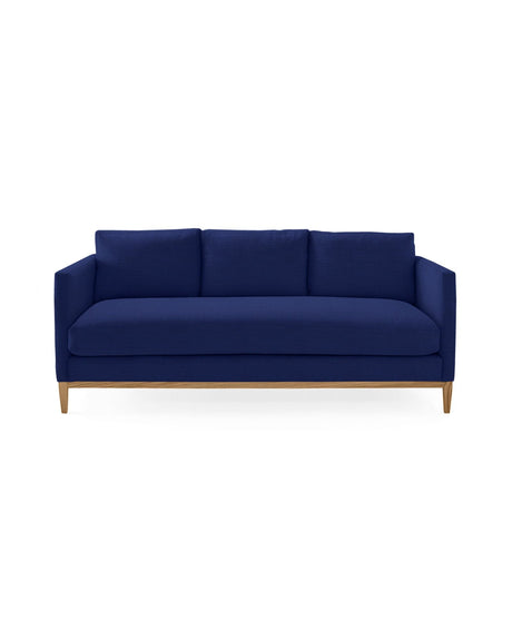 The Barton Sofa features a modern design with deep blue upholstery, clean lines, and wooden legs. It is a three-seater with a minimalistic style, complemented by three plush cushions on the backrest and set against a plain white background.