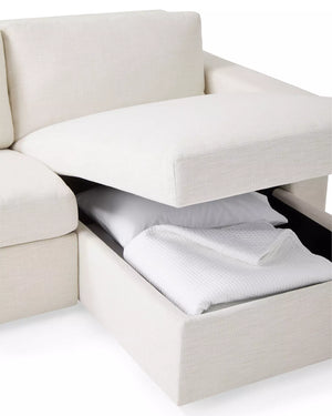 The Conover Sleeper Chaise Sectional in beige features a storage compartment beneath the seat cushion, revealing a folded white blanket and pillow that highlight its multifunctional design.