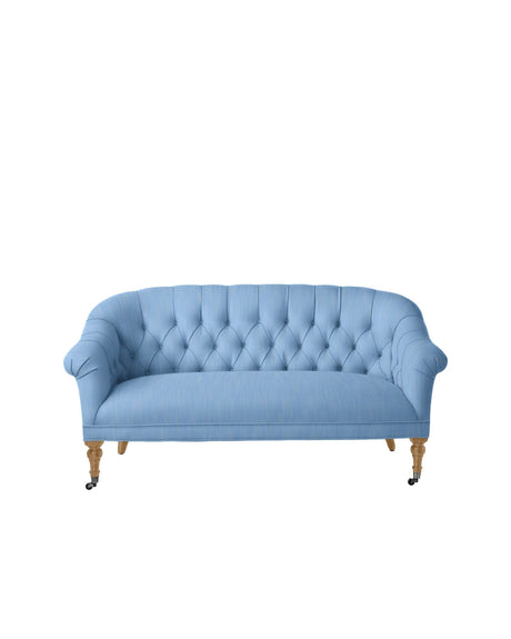 The Paxton Tufted Loveseat, featuring a light blue upholstery with curved arms and a wooden frame, stands on caster wheels. It boasts a classic design with three cushioned seats and is showcased against a plain white background.
