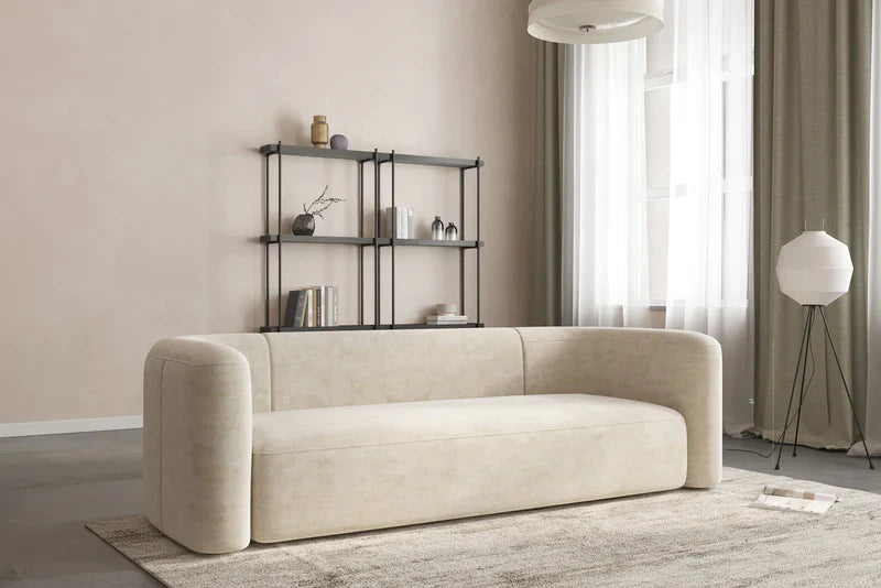 A minimalist living room features the Mellow Sectional Sofa by Acanva in beige, resting on a light-colored rug. Nearby, a black metal shelf holds books and decor against the wall, while a white floor lamp stands close by. Natural light filters through sheer curtains covering the tall windows.