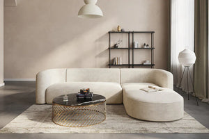 A modern living room features the Mellow Sectional Sofa by Acanva, with its elegant curves and beige upholstery. Complementing the sofa is a round glass coffee table encased in a gold frame, placed atop a textured rug. A black wall shelf and floor lamp sit against the tan wall, while white curtains drape over a large window.