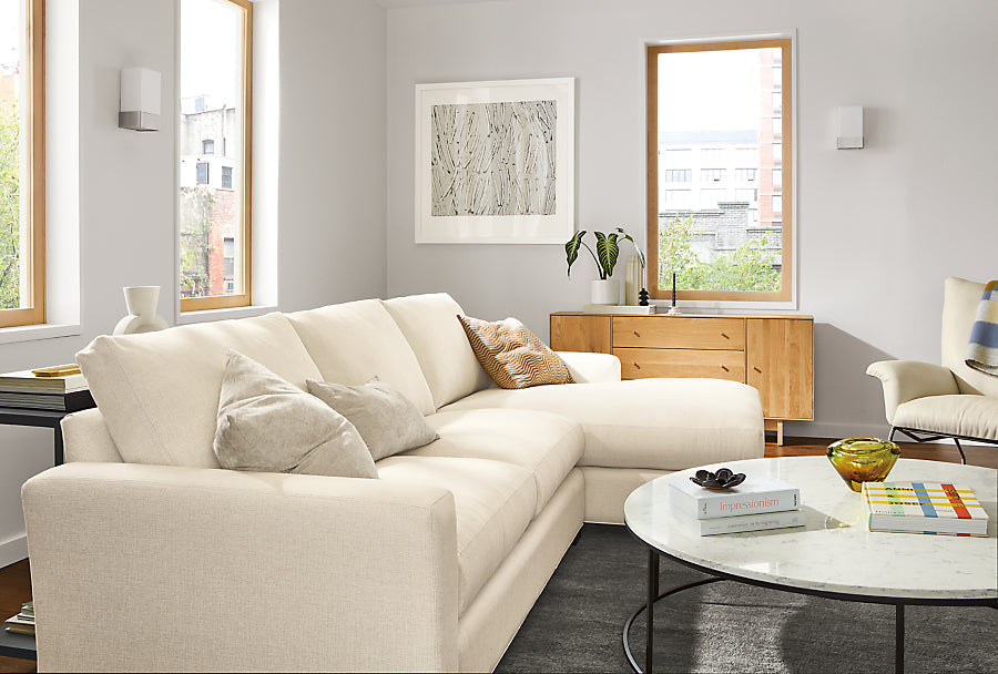 A modern living room features a spacious beige Metro Sofas with Chaise, complemented by a round marble coffee table and a wooden sideboard. The room boasts large windows, framed artwork on the walls, and various decorative items adorning the tables.